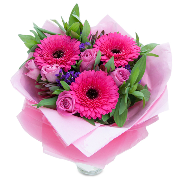 The bouquet reads like a small, joyful garden held together with care: three wide-eyed gerbera daisies in a vivid fuchsia sweep the gaze with their symmetrical petals and pronounced dark centres, while several soft lavender-pink roses sit nearby as demure, budding companions, their velvety petals still curved inward. Between these primary blooms, clusters of tiny purple statice lend a delicate, papery texture that twinkles against the larger forms, and generous sprays of deep green foliage - salal or eucalyptus - create a fresh, almost dewy backdrop that lifts the pinks visually and aromatically. Presentation is considered and graceful: soft, matte pale-pink tissue paper cradles the stems and is topped by a layer of sheer, slightly crinkled fuchsia-pink cellophane that catches shop light and gives the bouquet a subtle, celebratory gleam. The lower section reveals the top rim of a clear glass vase, ready for water and immediate display on a mantle, kitchen island, or café table. The scent is gentle - classic rose perfume warmed by a faint green note from the foliage - and the tactile experience ranges from the velvet softness of rose petals to the crisp resistance of the greens and the dry snap of statice. As a neighbourhood florist who knows Knightsbridge, I imagine this arrangement brightening a Chelsea street-party window, a quiet anniversary dinner near Harrods, or a bedside on a fresh morning after delivery from a local shop, carrying a message of affection, celebration, or simple encouragement.